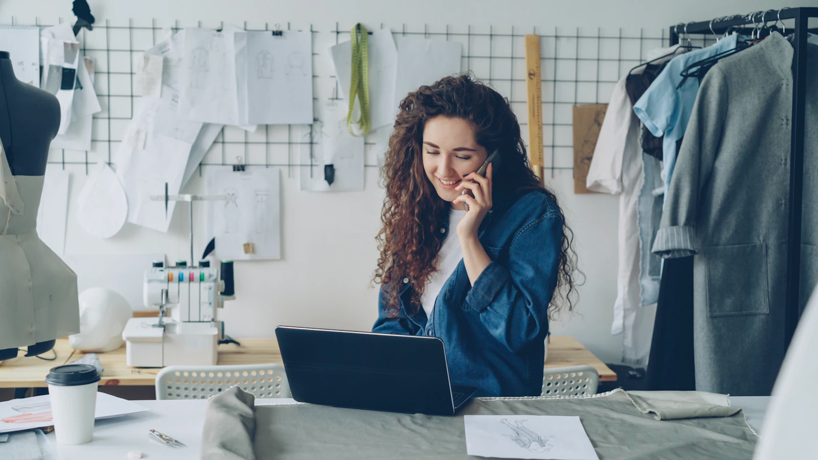 A shop owner on the phone at her laptop, between sewing patterns and a rail of clothes